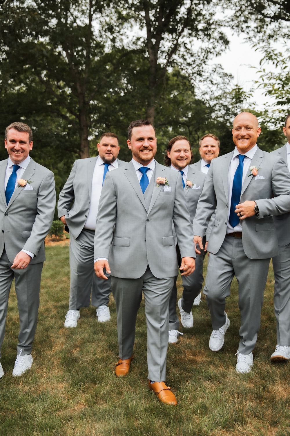 Groom and groomsmen in matching gray suits with blue ties and boutonnieres posing outdoors at a wedding.