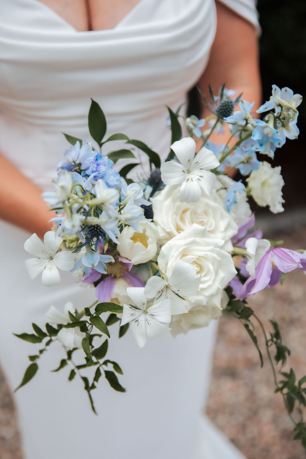 Bride holding a romantic bouquet of white roses, blue delphiniums, and purple flowers with greenery.