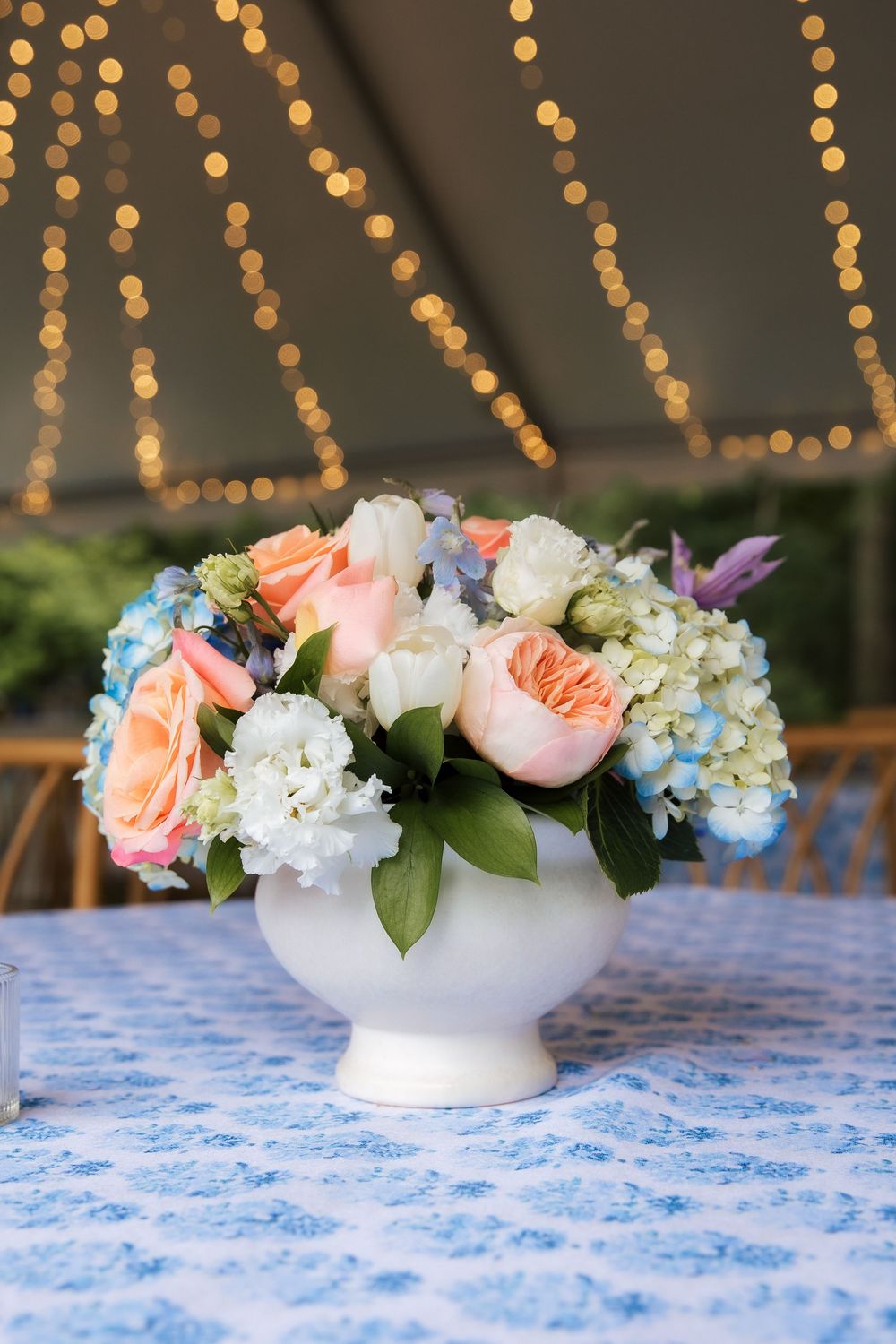 White bowl vase with peach roses, hydrangeas, and mixed blooms on blue floral tablecloth under warm bokeh string lights.