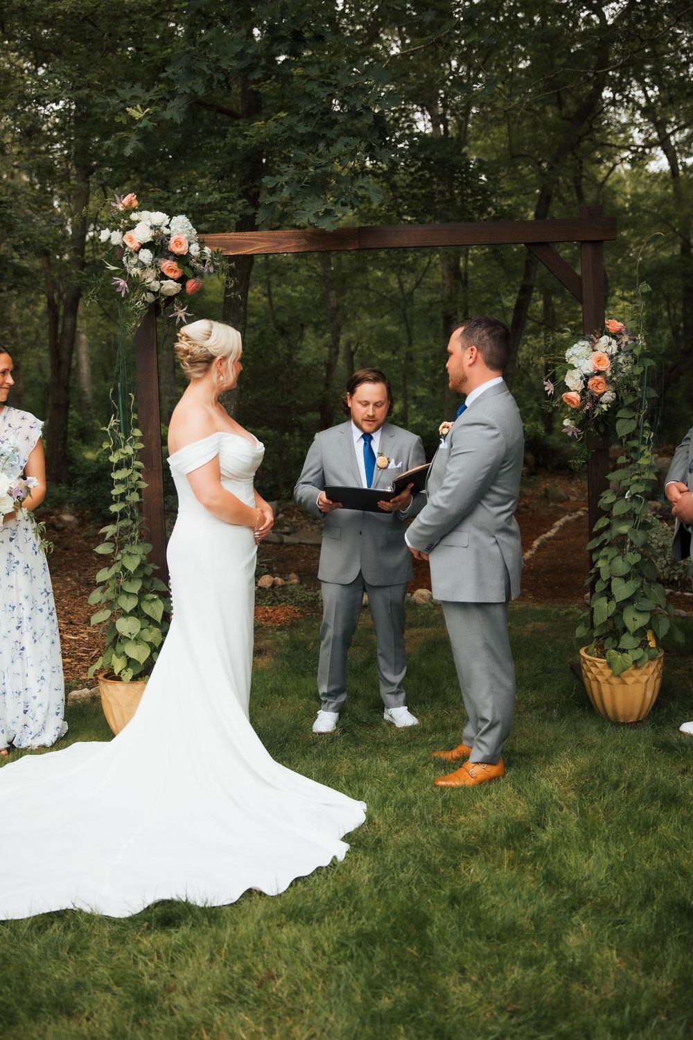 Outdoor wedding ceremony under wooden arch with floral arrangements, bride in white gown facing groom in gray suit with officiant.
