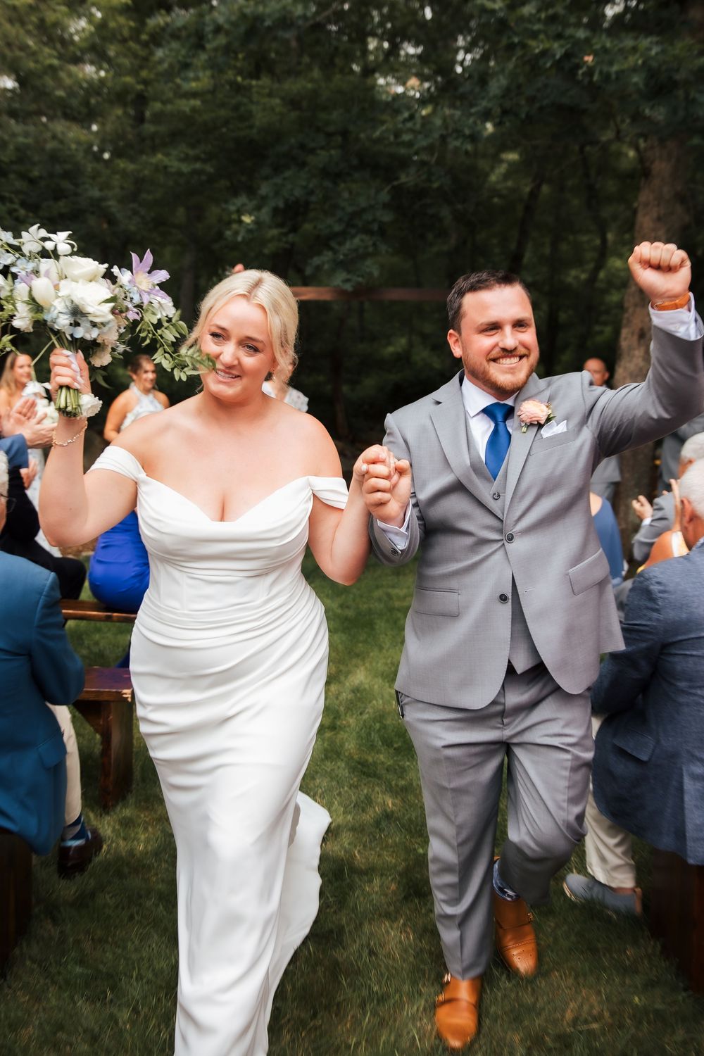 Joyful newlyweds walk down outdoor aisle, bride in white gown holding bouquet, groom in gray suit raising fist.