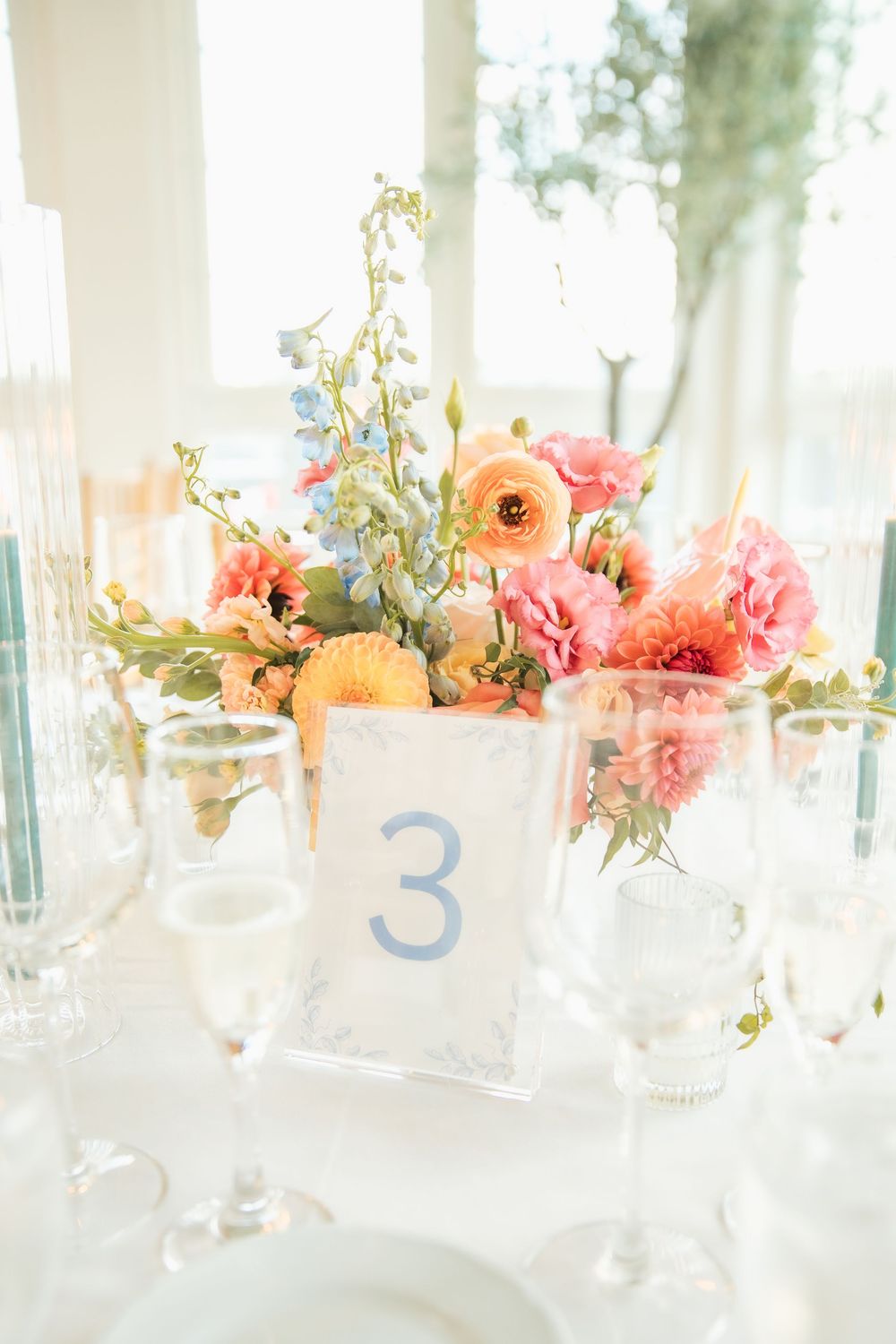 Elegant wedding table centerpiece with coral, peach, and blue flowers beside a number 3 table card and crystal glassware.