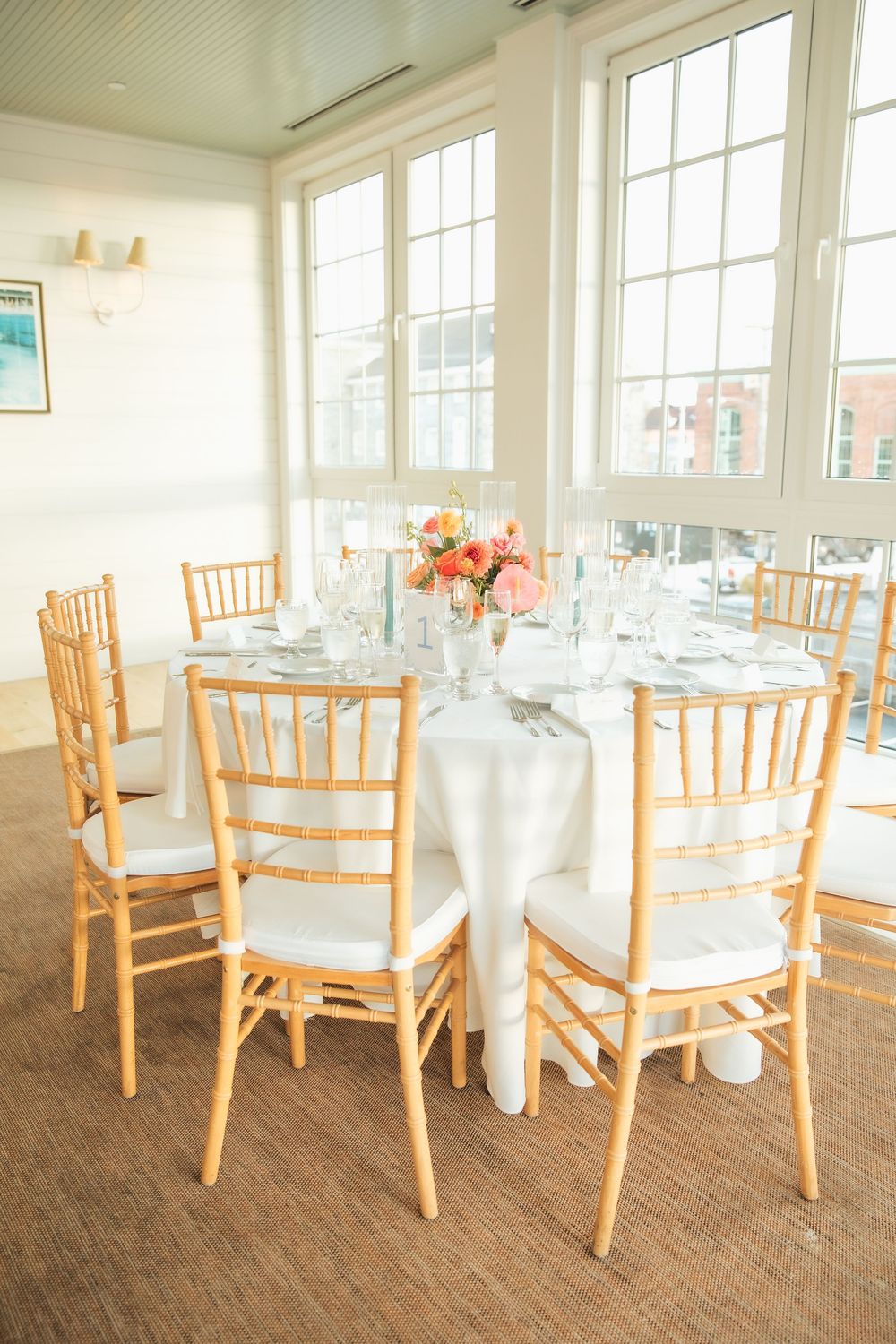 Elegant wedding reception table with gold chiavari chairs, white linens, and pink floral centerpiece in a bright venue.
