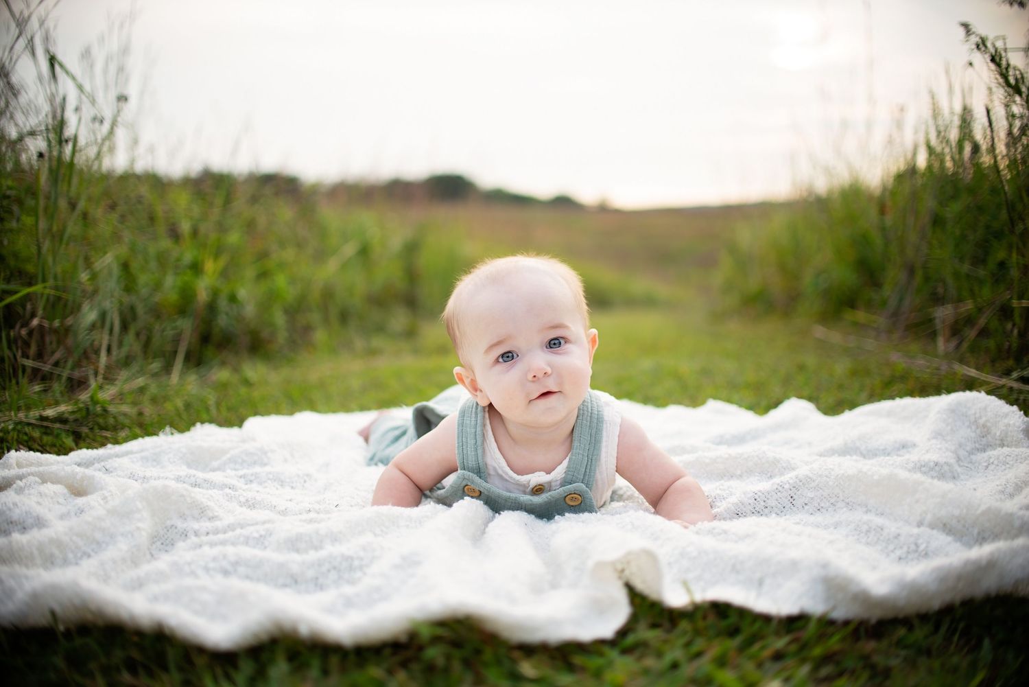 Adorable baby in a mint romper lies on a white blanket in a lush green field.