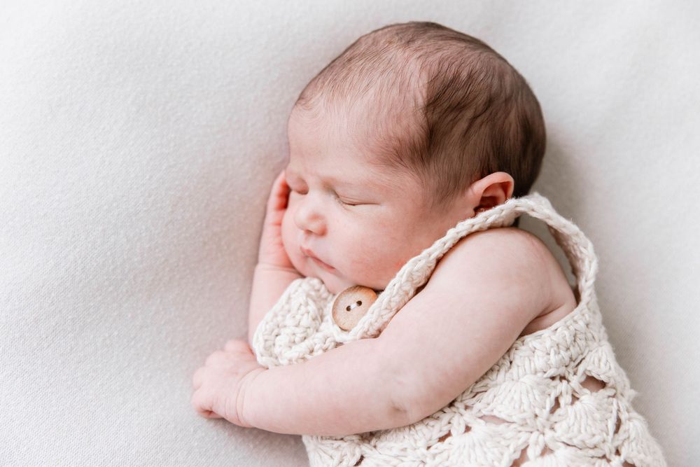 Sleeping baby wearing white lace dress lying on soft white blanket in peaceful newborn photography pose.