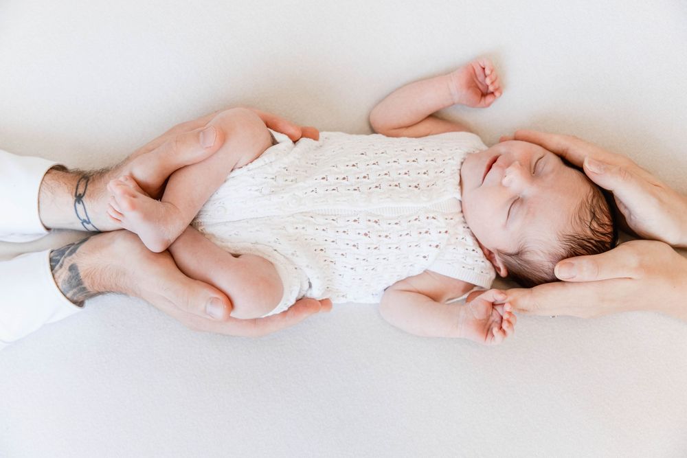A sweet baby wearing a white lace dress lies peacefully on a white background while being gently cradled by hands.