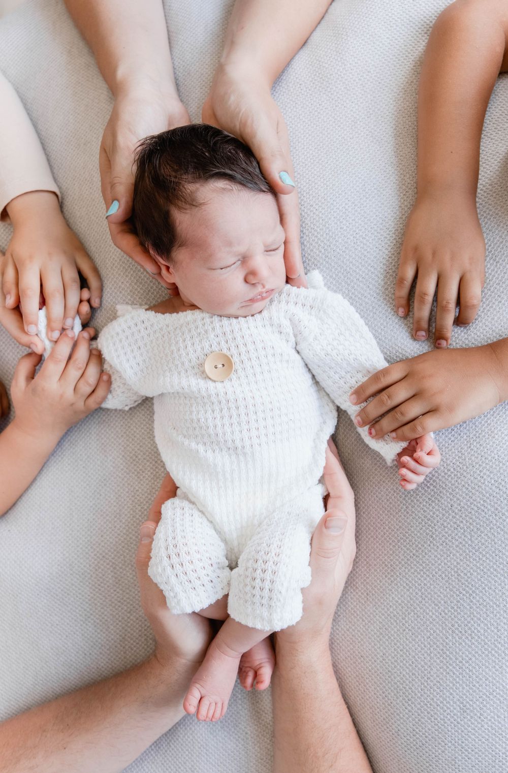 Newborn baby wearing white knit romper rests on gray fabric while small hands reach to touch them.