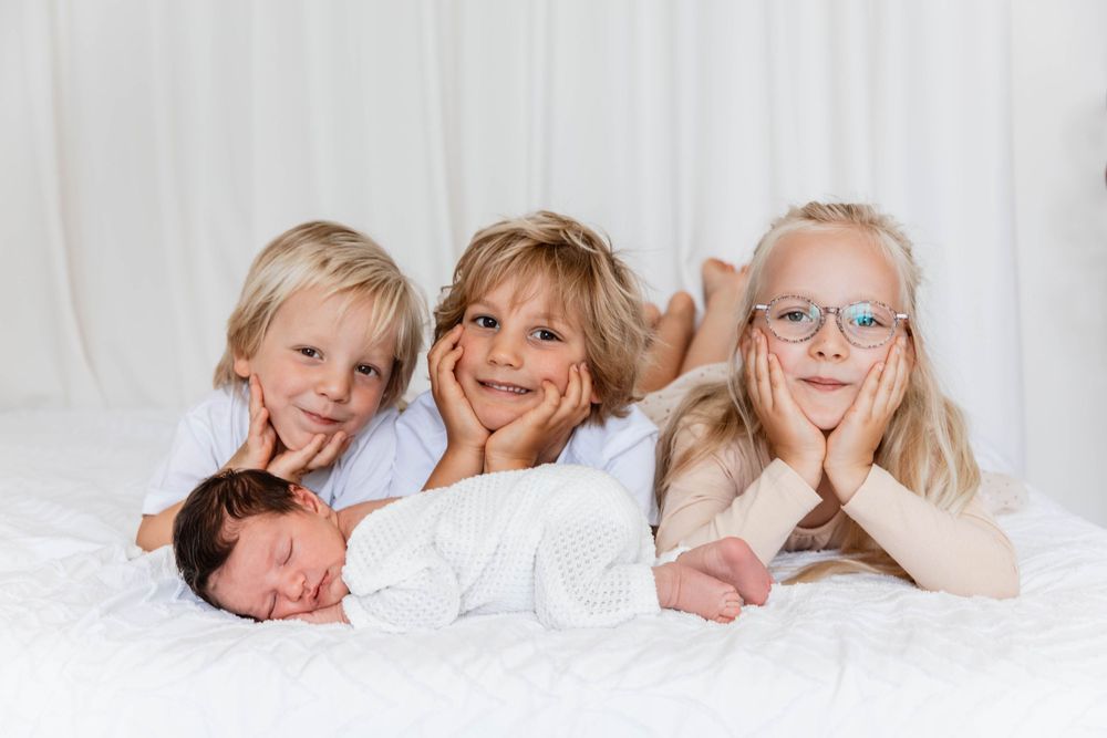 Four siblings lying together on white bedding while posing and smiling for a portrait.
