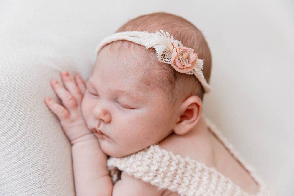 A sleeping infant wearing a delicate pink floral headband and knit top rests peacefully on white bedding.