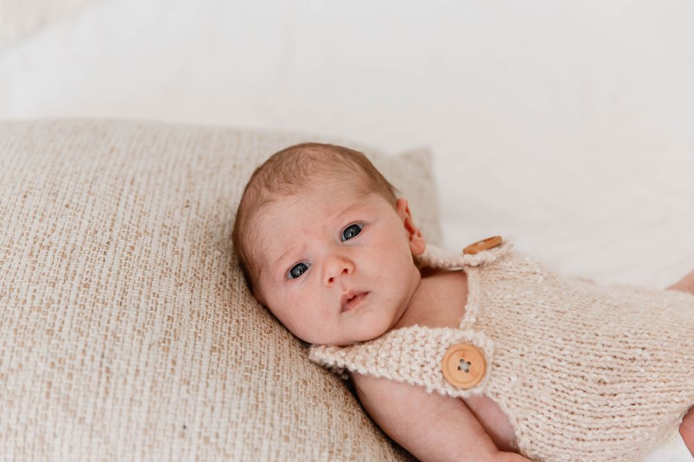 A baby in a cream-colored knit outfit lies on a textured beige blanket with a wooden button detail.