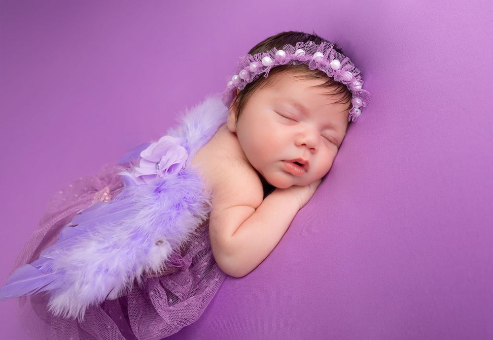 Sleeping newborn wearing purple flower crown and wings on purple background in artistic portrait.