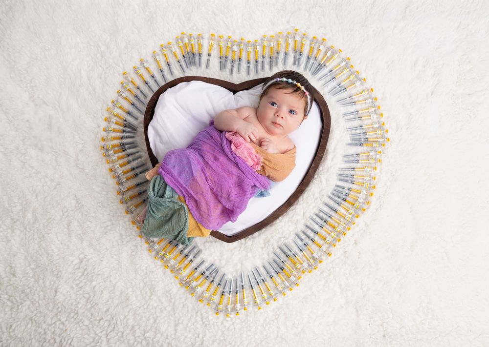 Newborn sleeping in heart-shaped basket surrounded by safety pins and soft fabrics.