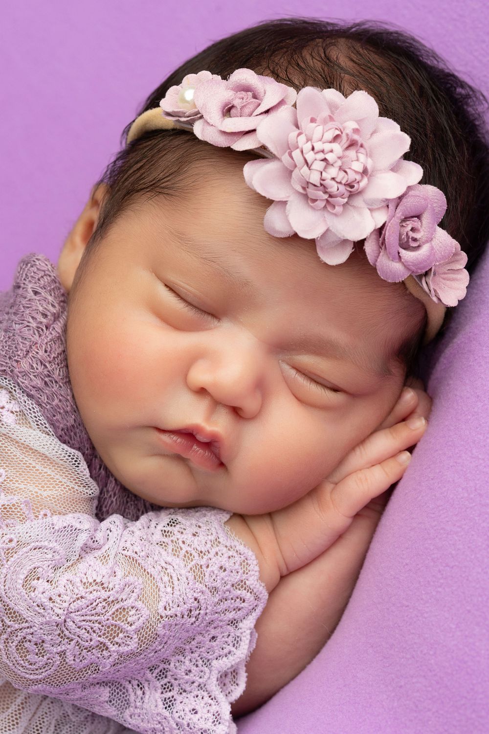 Sleeping infant with pink floral headband wearing lace dress against purple backdrop.