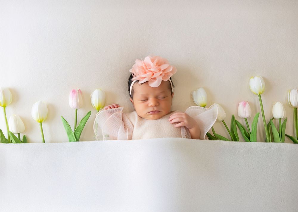 Newborn in pink dress and floral headband lies peacefully among white tulips in spring-themed portrait setting.