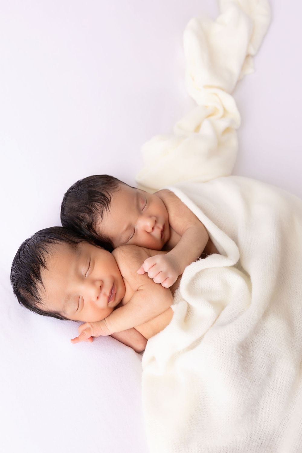 Two sleeping newborn babies wrapped in soft white blanket laying close together on white background.