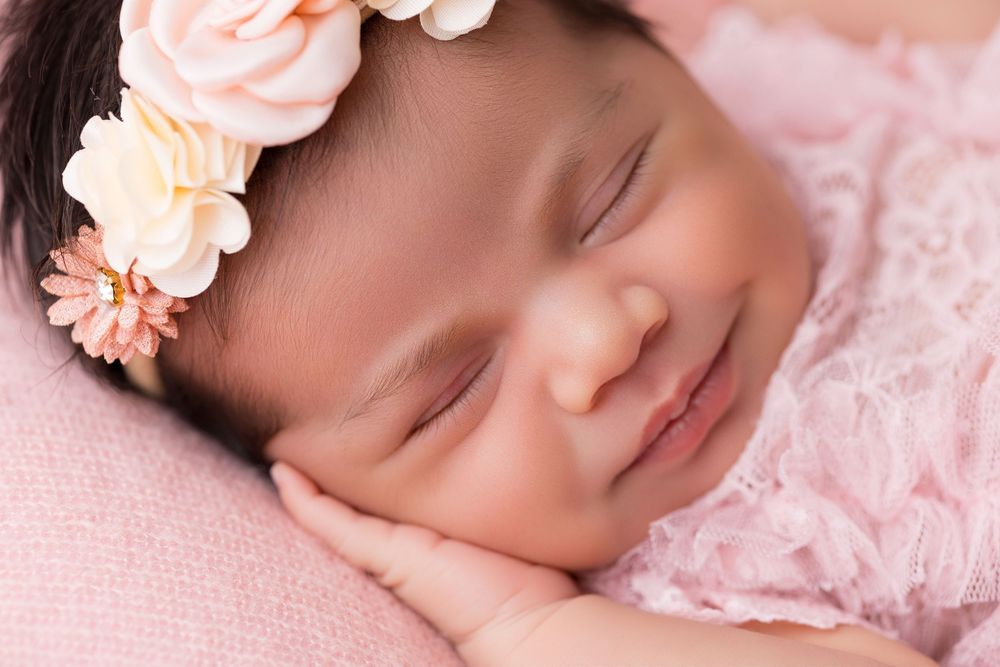Smiling newborn baby girl sleeping peacefully wearing a pink floral headband and lace outfit on a soft pink blanket.