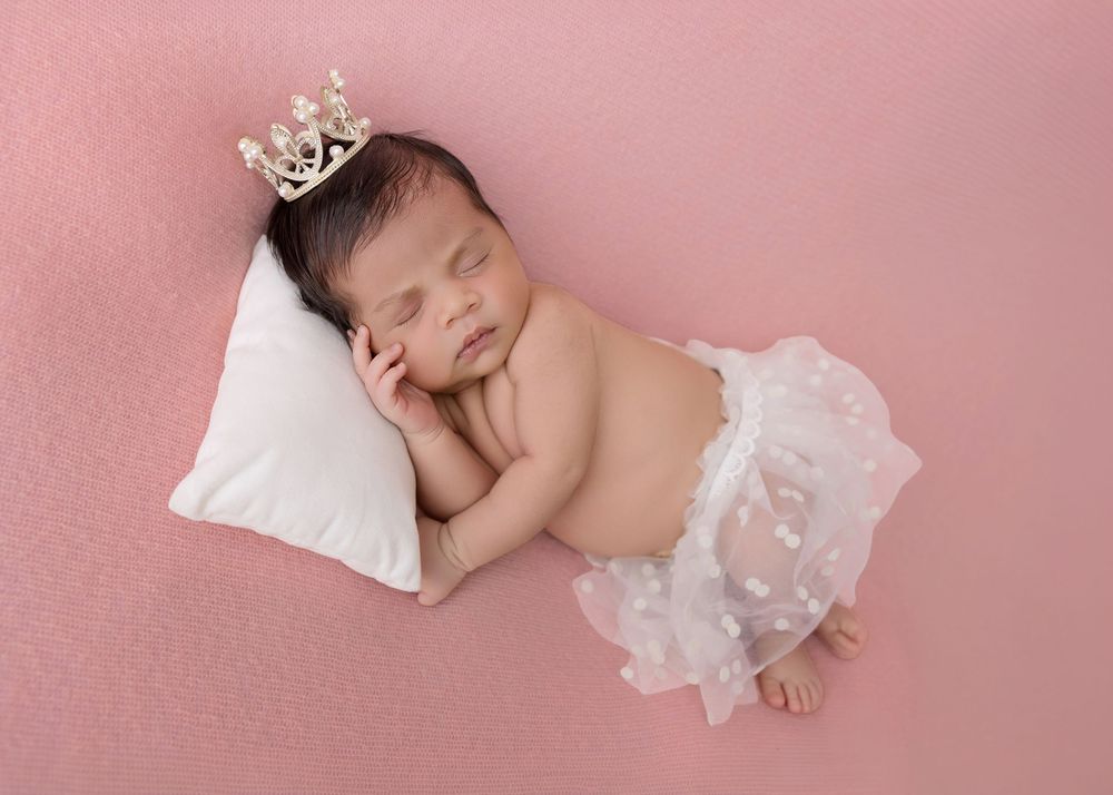 Newborn baby girl sleeping on white pillow wearing tiny crown and white tutu skirt on pink background.