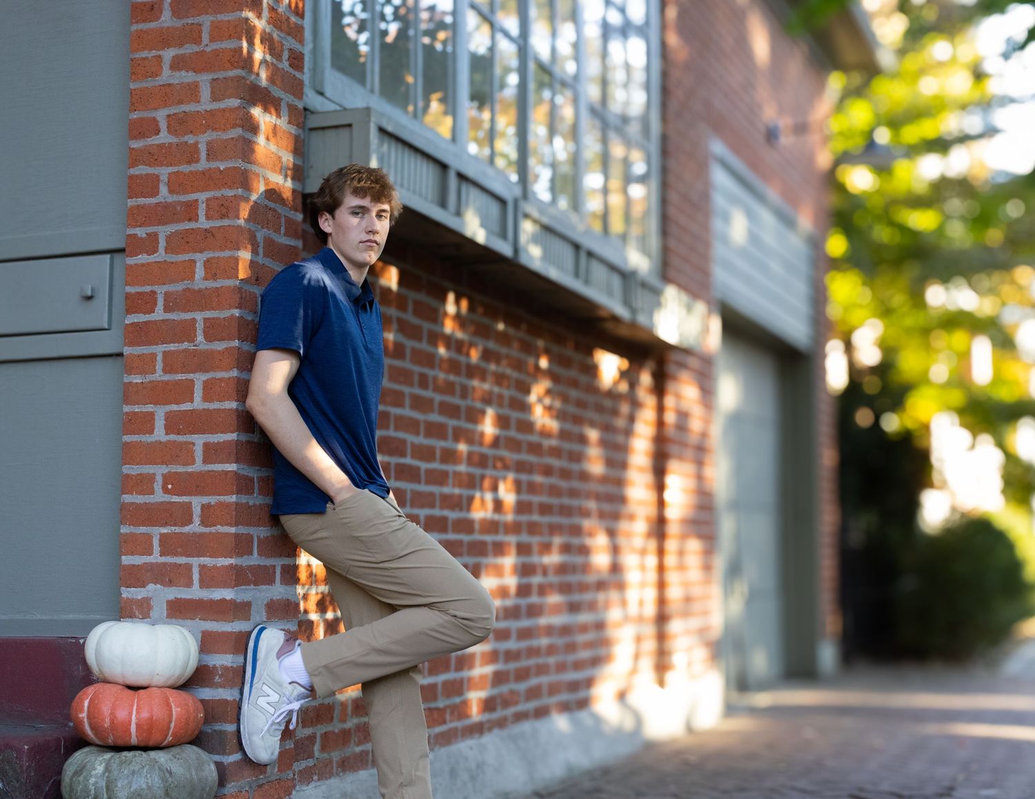 Young man in blue polo leaning against red brick wall with pumpkins in autumn setting.