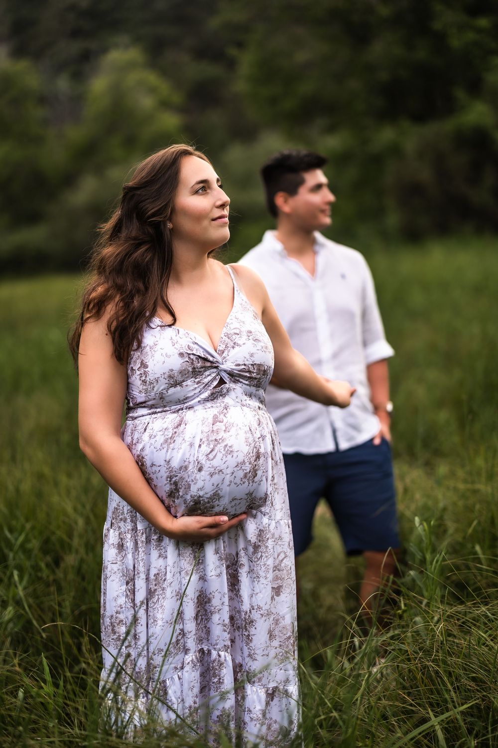 Couple poses in grassy field as woman in flowing white dress cradles her pregnant belly during outdoor photoshoot.