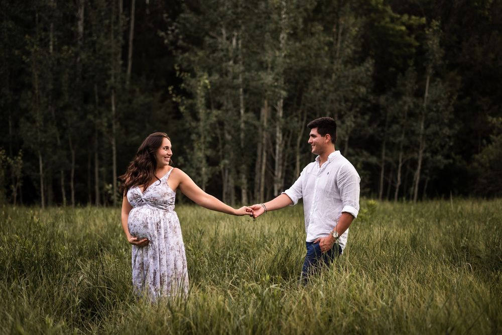 A couple holds hands in a tall grass meadow surrounded by green forest, the woman wearing a flowing white dress.