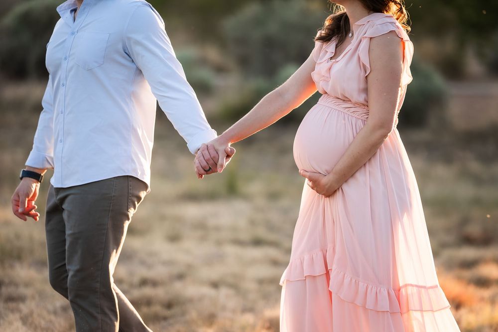 Pregnant couple holding hands outdoors in golden light, woman in pink dress cradling baby bump.