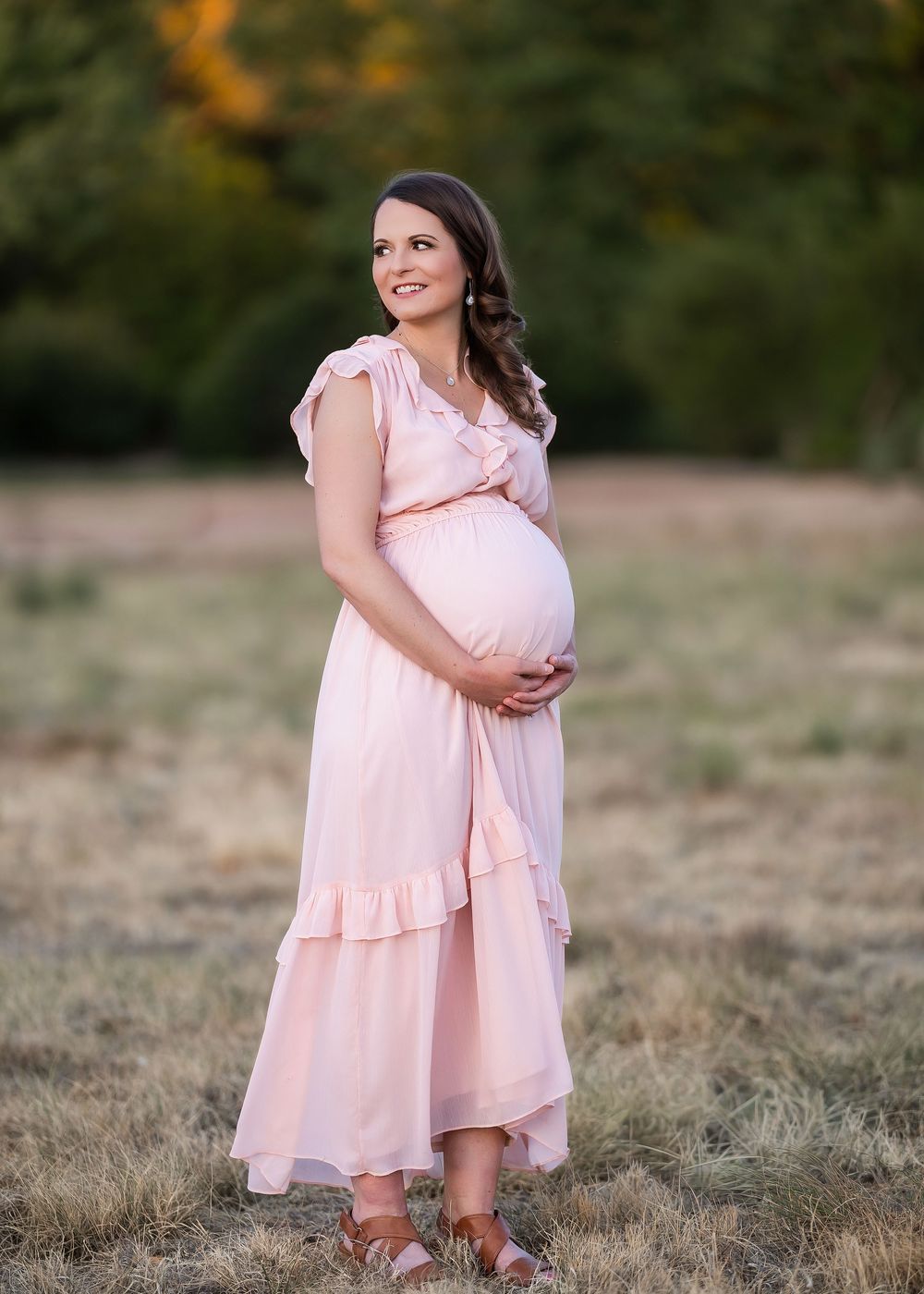 Pregnant woman in pink maxi dress cradling baby bump outdoors in golden hour light during maternity photoshoot.