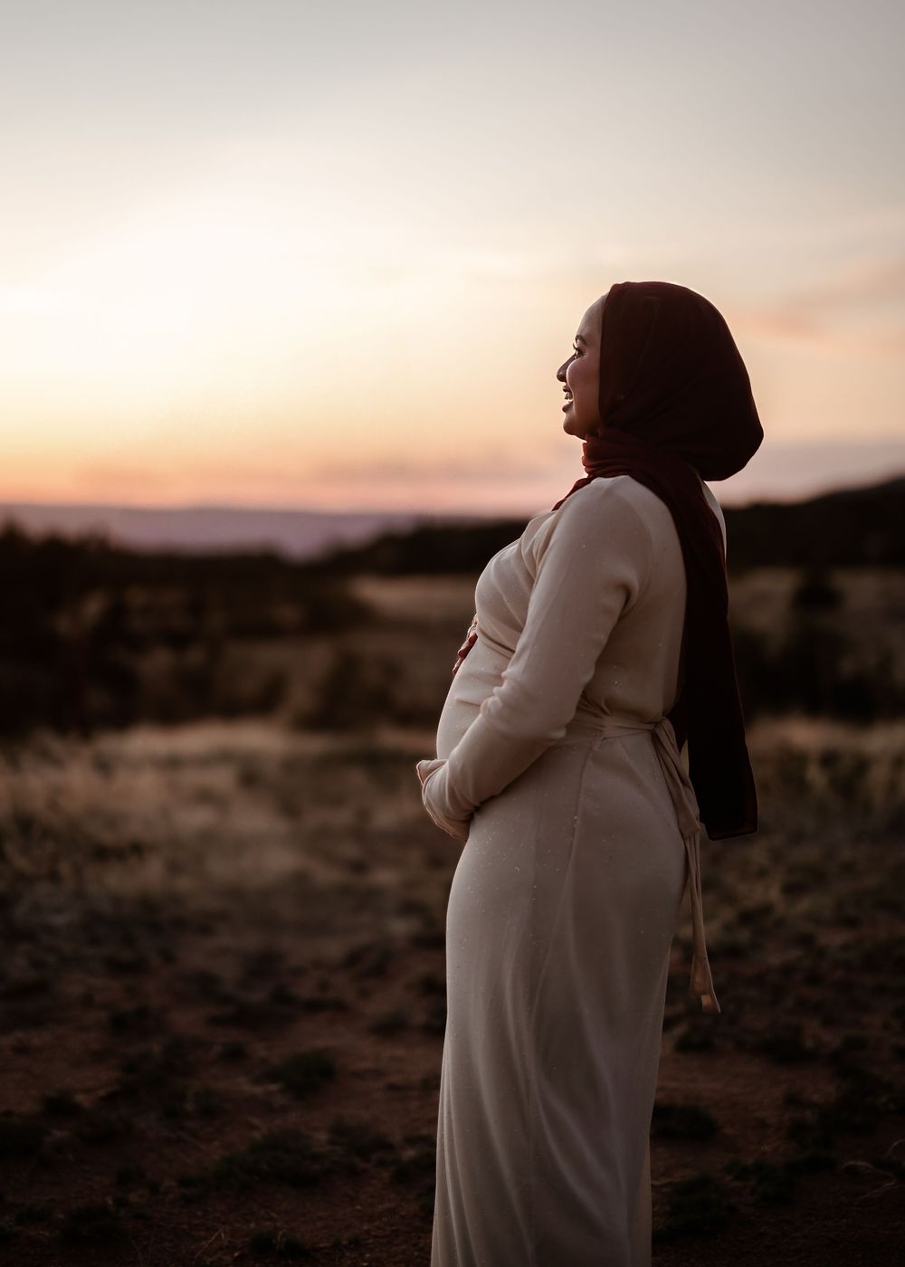 Silhouetted figure in a white dress stands contemplatively against a warm sunset sky in a desert landscape.