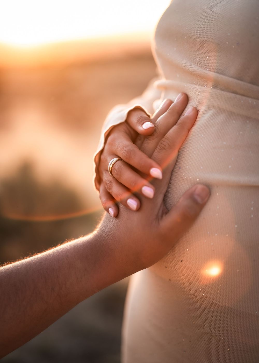Gentle hands touch a pregnant belly during golden sunset, creating an intimate maternity silhouette.