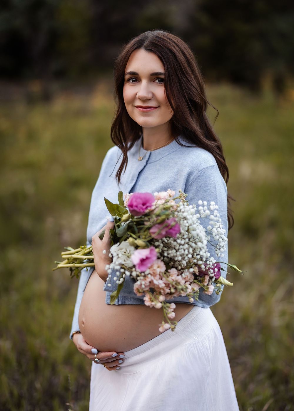 Pregnant woman in blue cardigan holding a bouquet of pink and white wildflowers outdoors in a field.