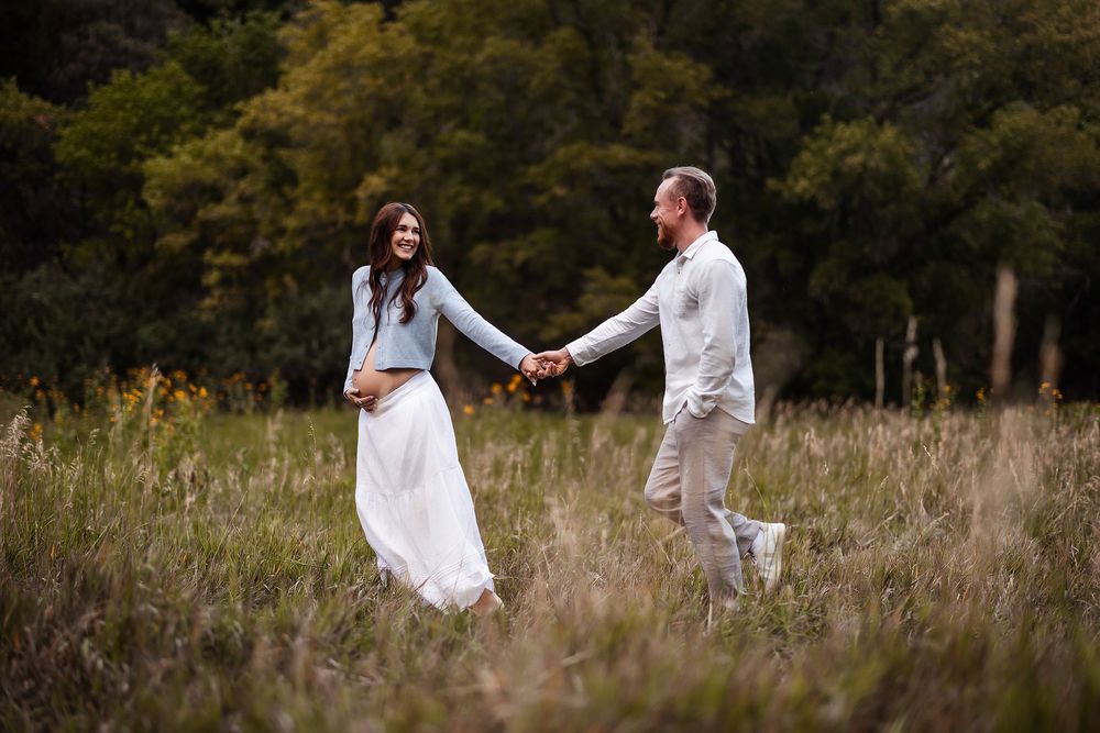 Couple holding hands and smiling in a lush green meadow, dressed in white and blue for an outdoor engagement session.