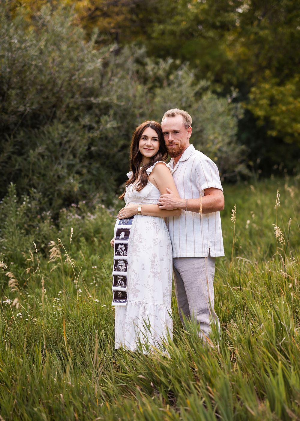 Couple embracing in a field, woman in white dress holding ultrasound photos, announcing pregnancy outdoors.