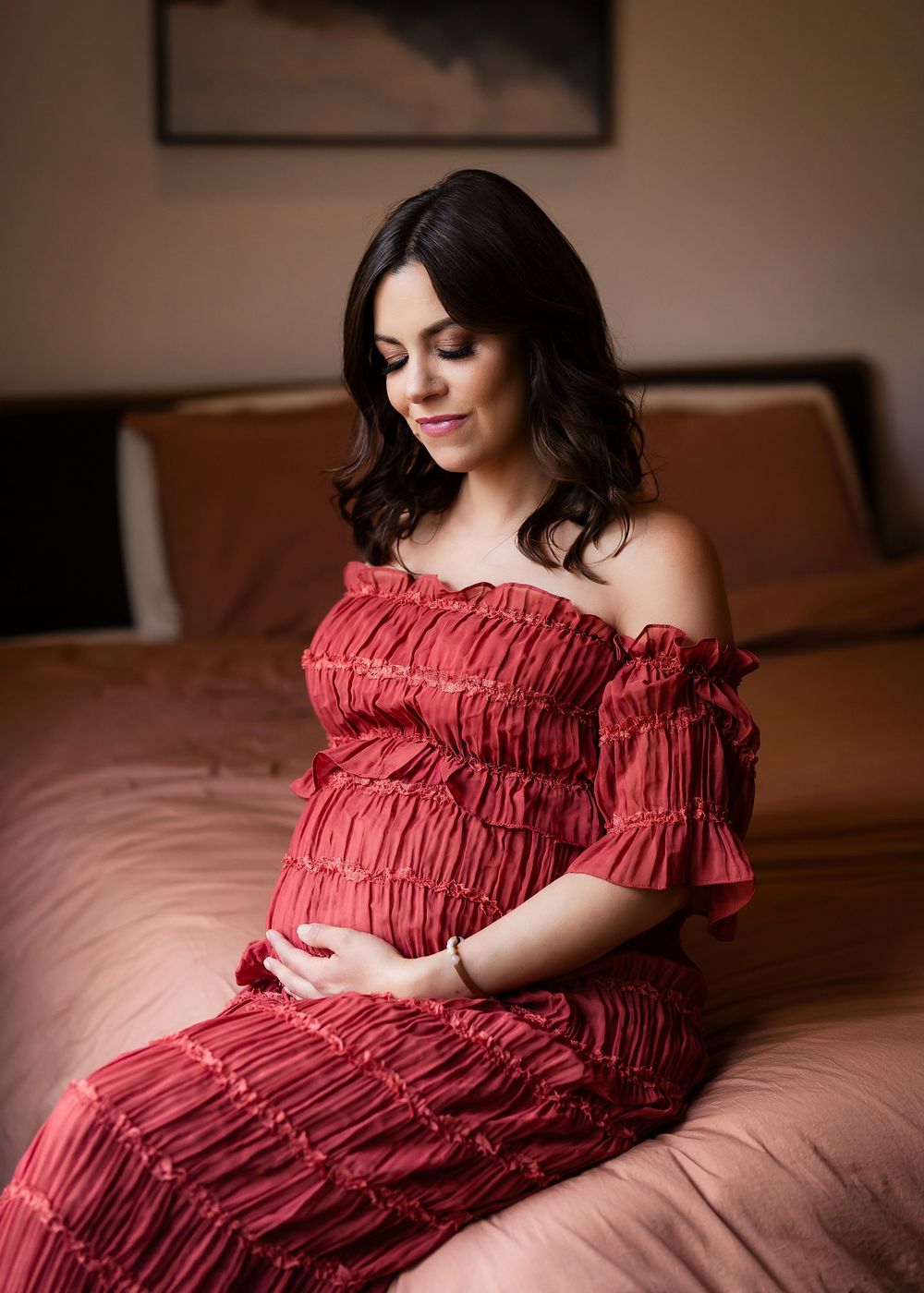Pregnant woman in red ruffled off-shoulder dress sitting on bed, cradling baby bump, maternity photo session.