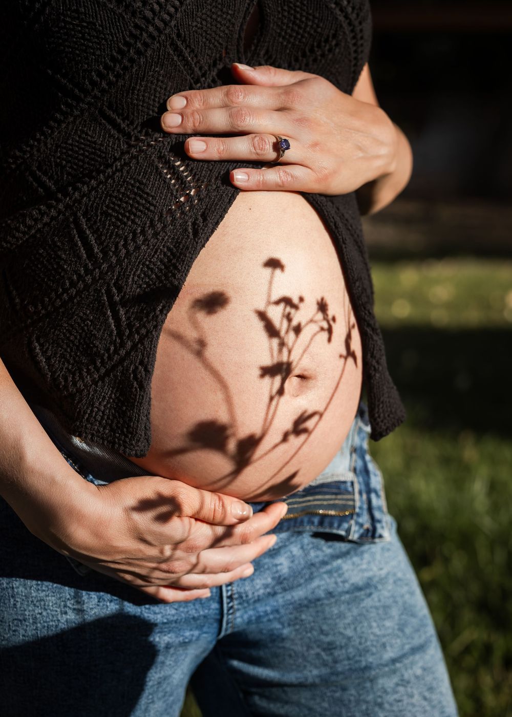 Pregnant woman in jeans cradling bare baby bump with floral shadows cast across skin in natural sunlight.