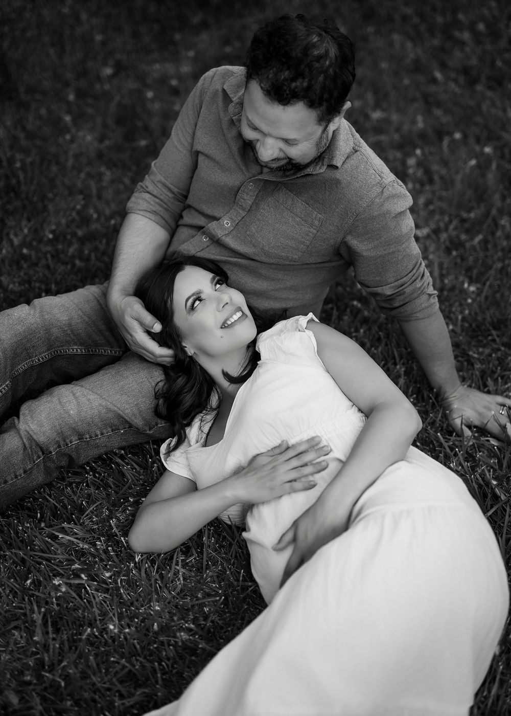 Black and white photo of a couple relaxing on grass, woman in white dress lying down smiling up at man beside her.