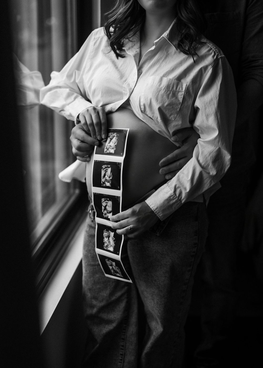 Black and white maternity photo of pregnant woman holding ultrasound strip near window, showing baby bump.