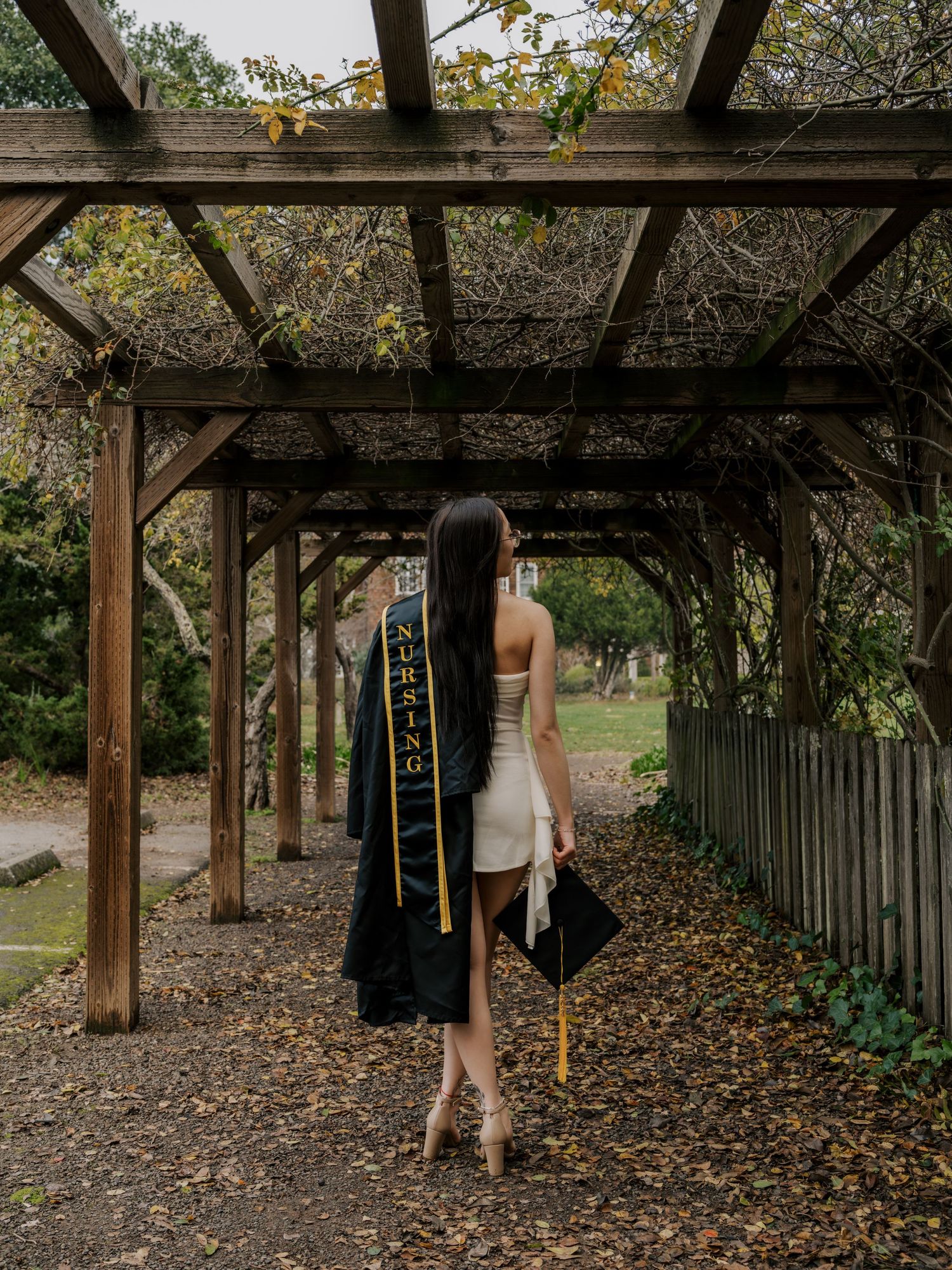 Graduate in a white dress holds graduation attire while walking through a wooden pergola covered in fall leaves.
