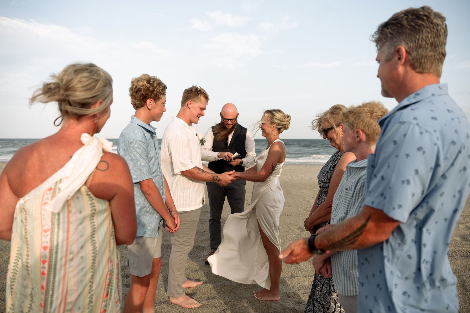 A small wedding ceremony takes place on a beach at sunset with guests gathered in a circle.