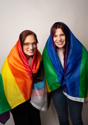 Two people wrapped in rainbow pride flags posing together against neutral background.