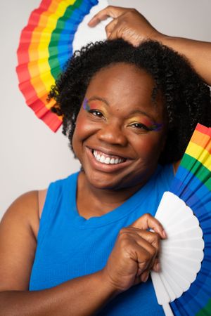 Cheerful poses with rainbow folding fan against light studio background.