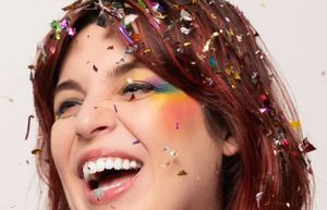 Closeup of someone with red hair surrounded by falling rainbow confetti and glitter.