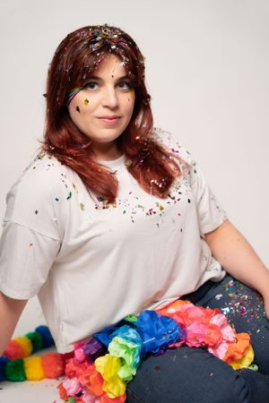 Person in white t-shirt posing with vibrant rainbow decorations and confetti.