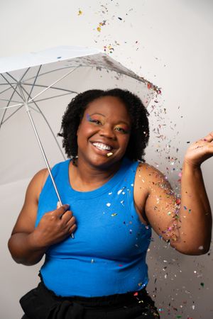 Person posing with white umbrella and colorful confetti in studio setting.