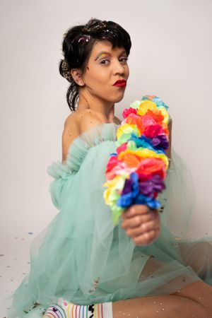 Person wearing a mint green dress poses with a rainbow-colored pom pom against a white backdrop.