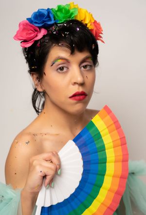 Model wearing floral crown and mint dress poses with rainbow fan in studio setting.