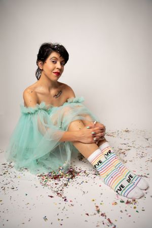 Model in mint tulle dress shows off pride-themed socks while lounging on confetti-covered floor.