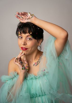 Model in mint green tulle dress poses playfully against grey backdrop with glitter accents.
