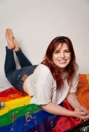 Person lying on rainbow pride flag with scattered glitter in studio setting.