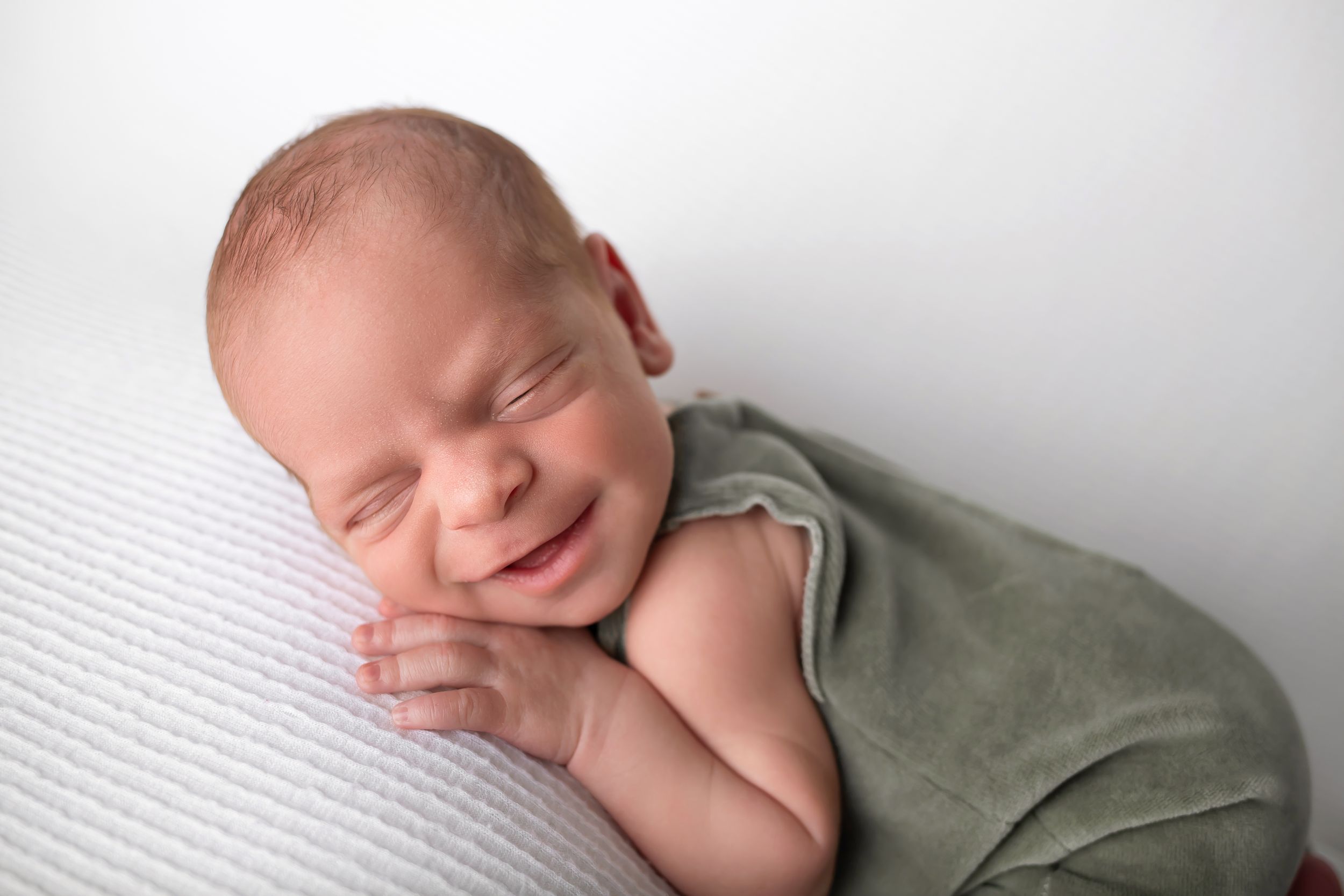 A sleeping newborn baby in gray outfit smiling while resting on white textured surface.