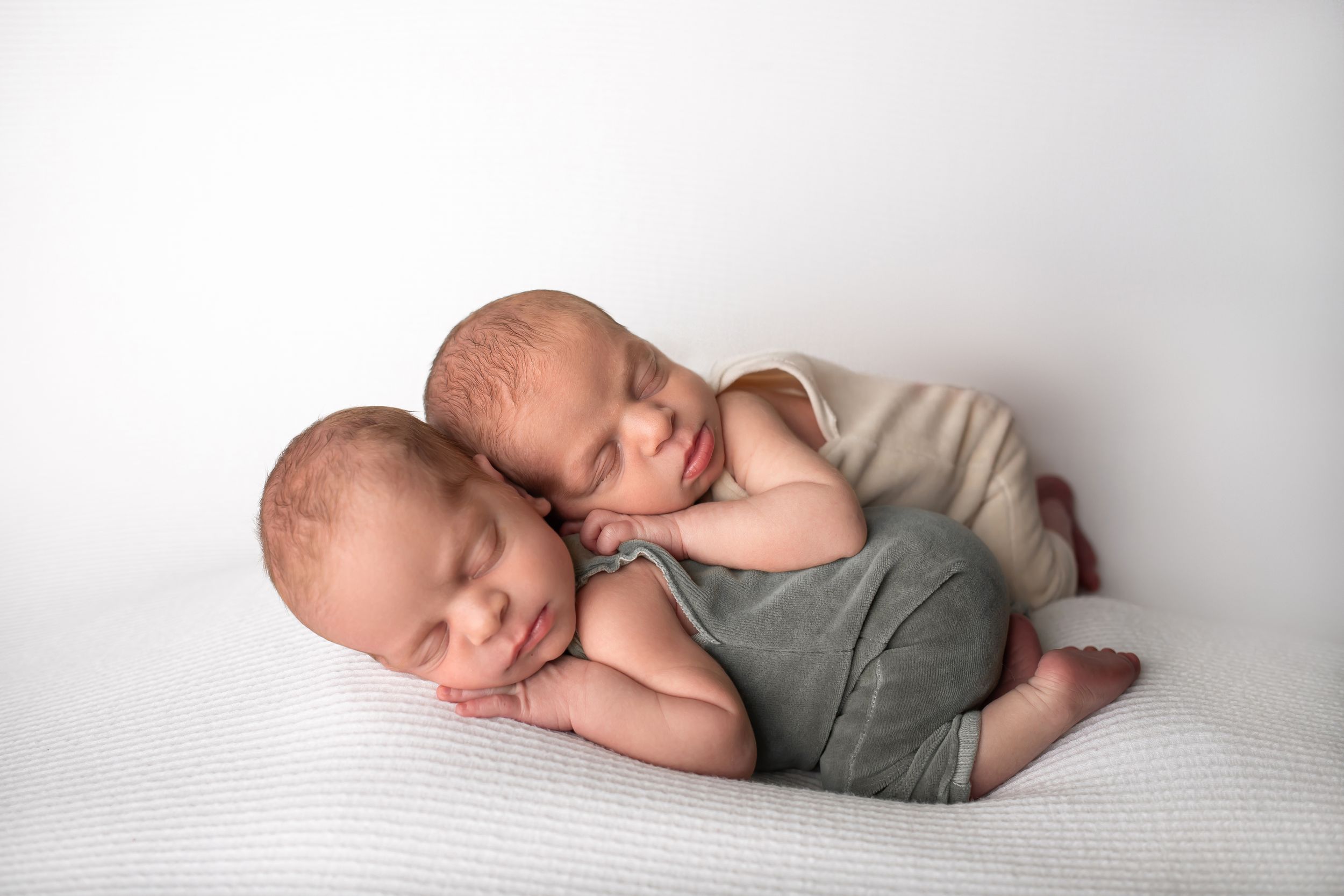 Two sleeping newborn babies in coordinating outfits cuddled together on white backdrop.