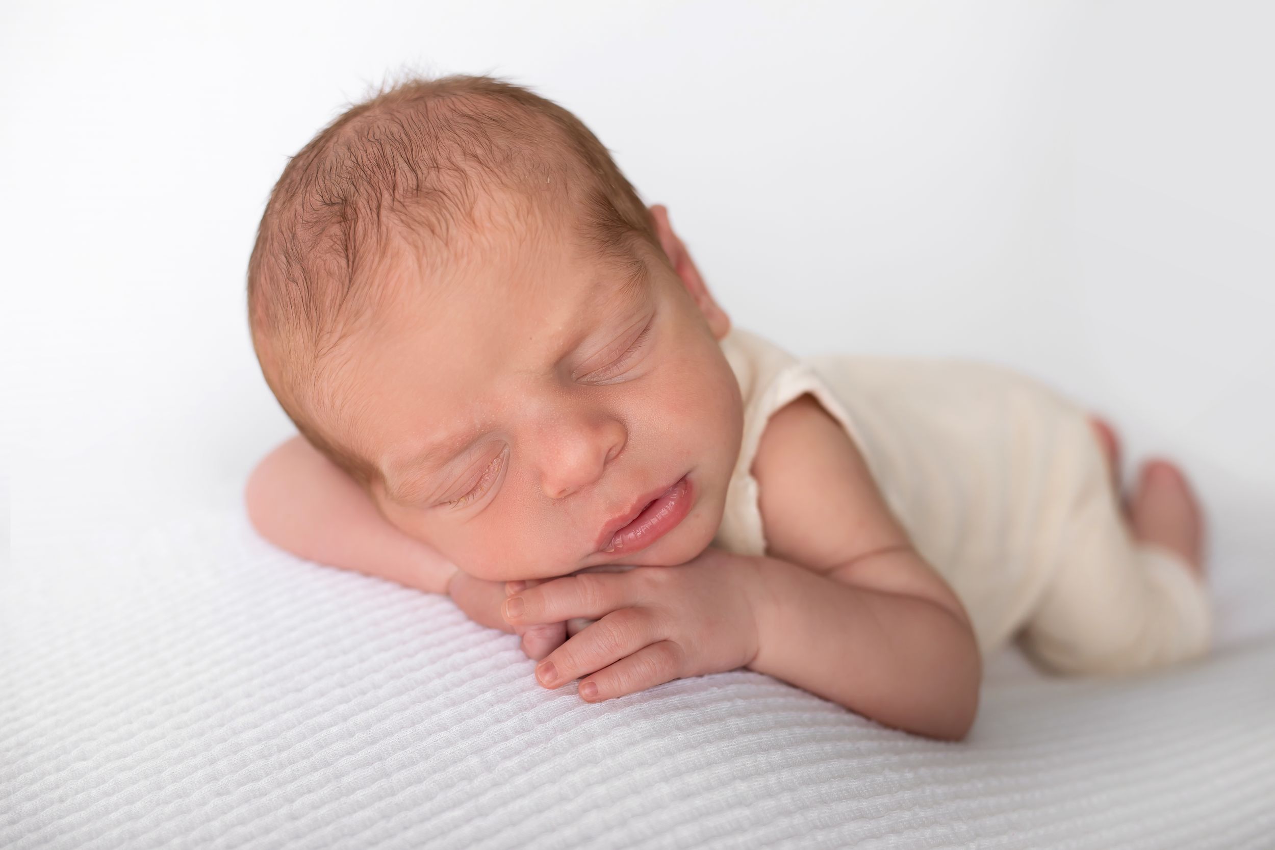 Peaceful newborn sleeping on white surface with hands tucked under chin.